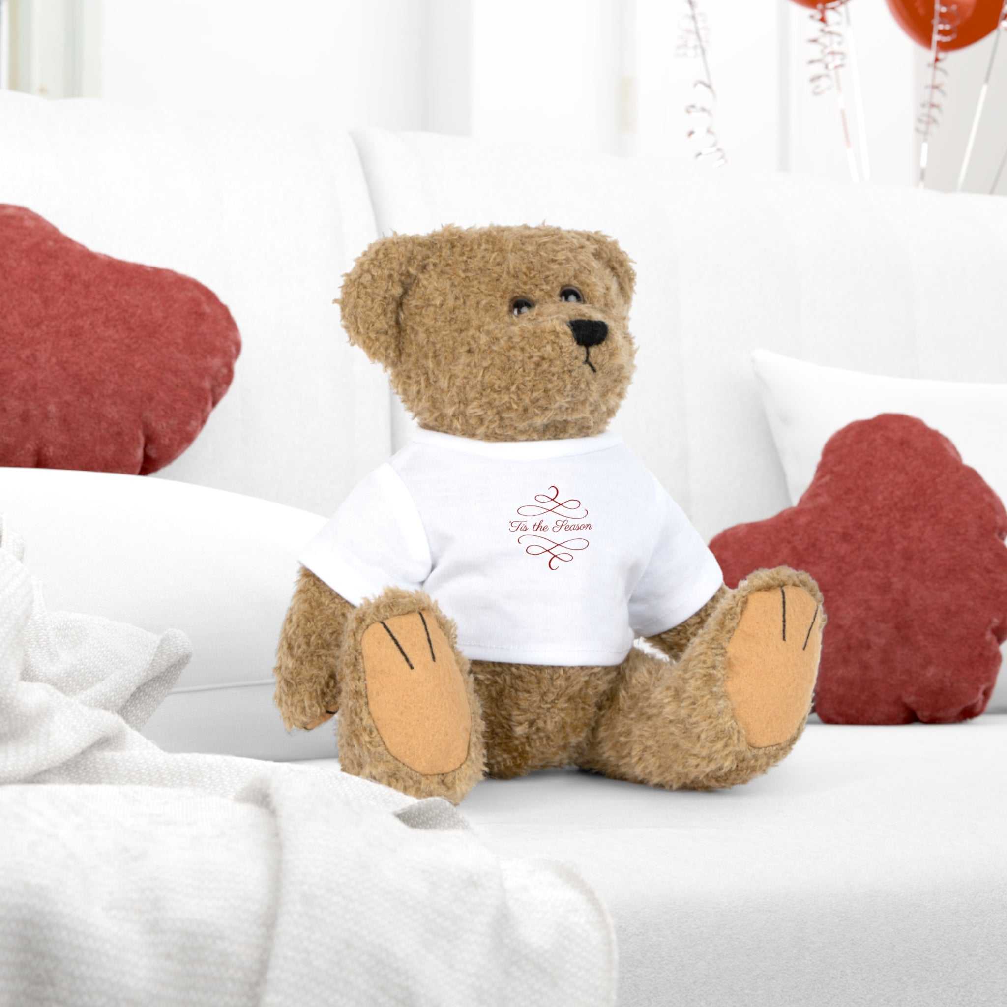 Tis the Season brown teddy bear plush toy wearing a removable holiday-themed white t-shirt sitting on a white couch with red pillows